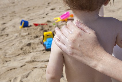 Mother applying sun cream to her baby on back with beach toys at bottom